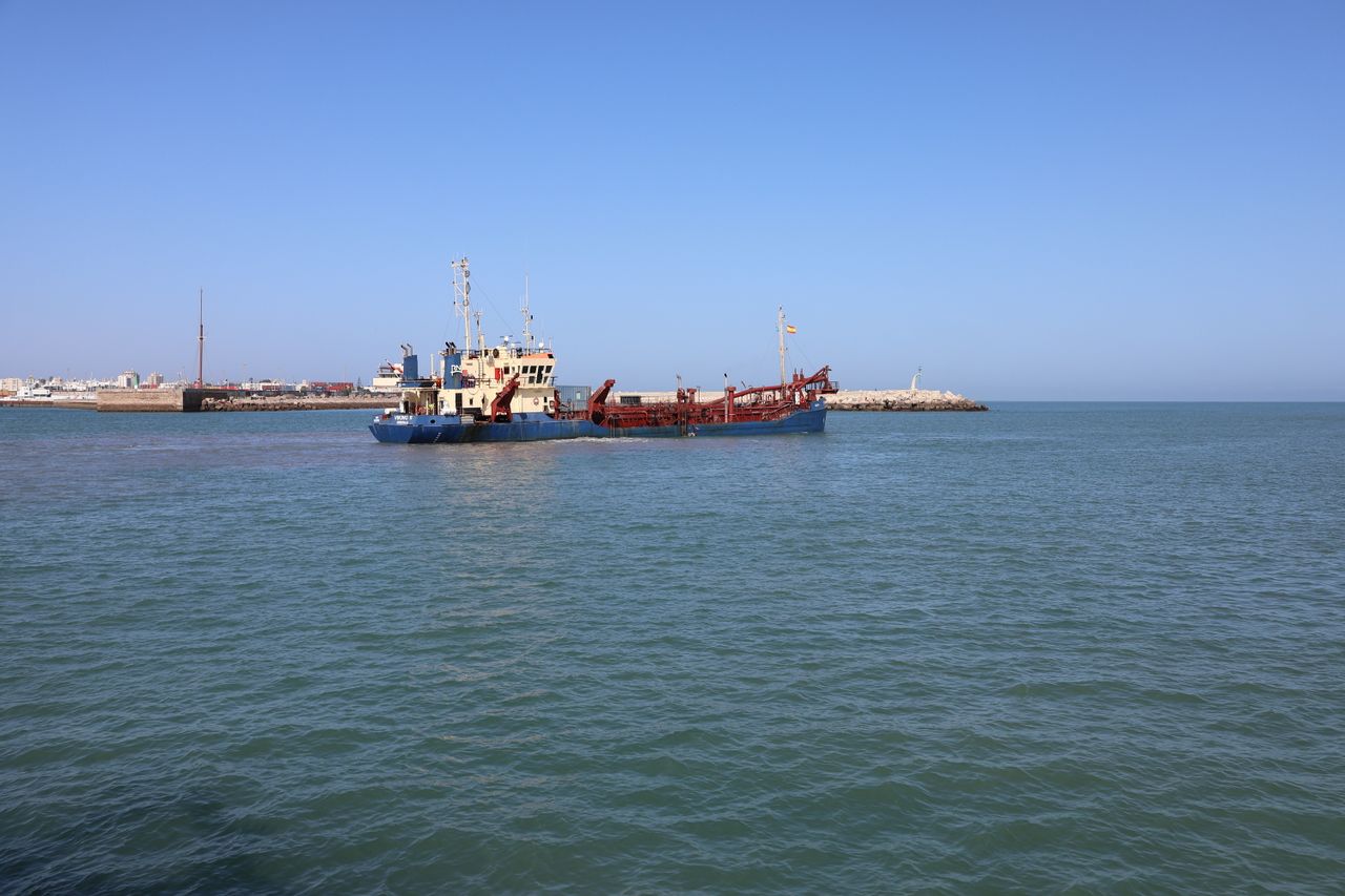 A dredger vessel operates in the scenic port of Cádiz, Spain under clear blue skies.